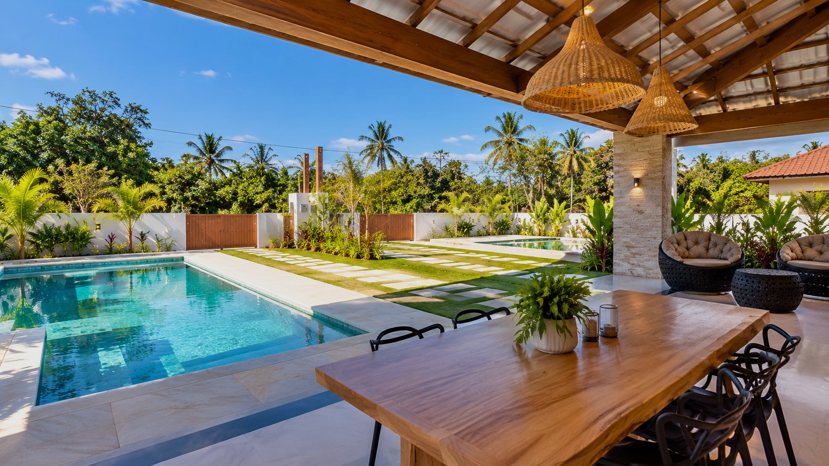 Überdachter Outdoor-Essbereich einer modernen Ferienimmobilie mit großzügigem Massivholztisch, privatem Pool und tropisch gestaltetem Garten.