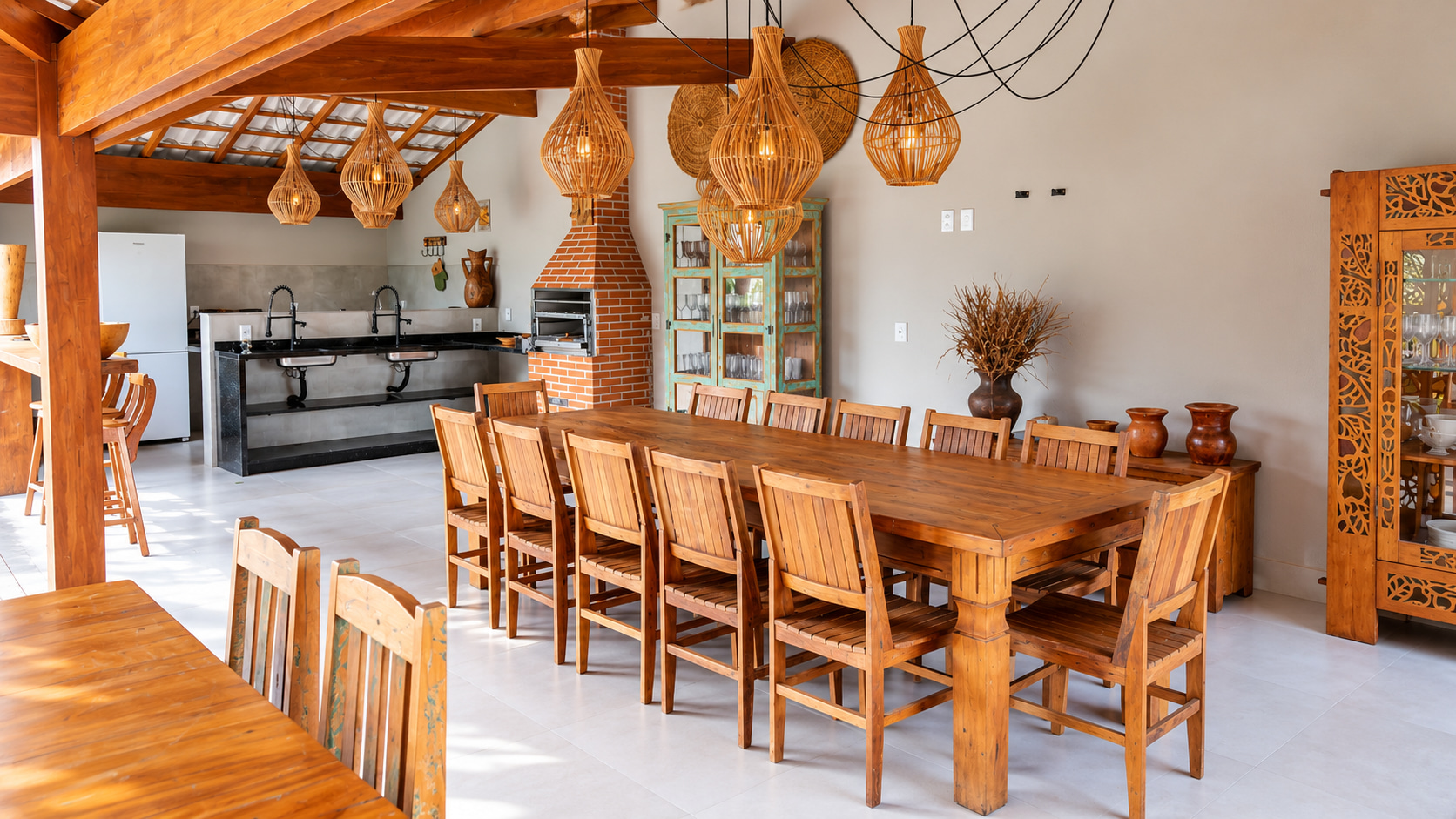 Covered communal dining area at a boutique guesthouse with a large wooden table, outdoor kitchen, built-in brick grill, and woven pendant lights.