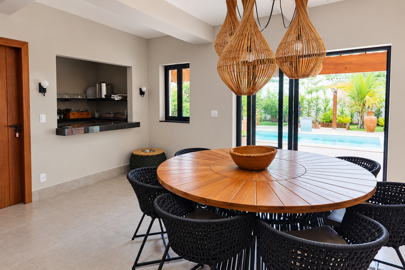 Bright dining area at a boutique hospitality property, featuring a round wooden table, woven pendant lights, kitchen pass-through, and pool views.