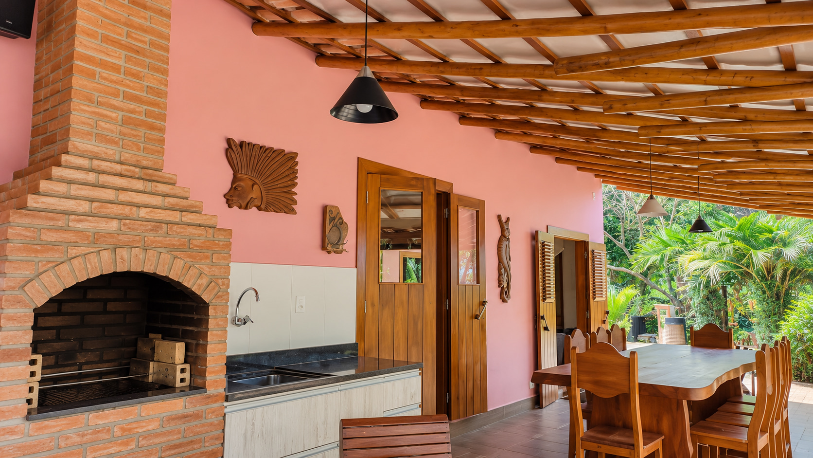 Überdachte Außenküche der Ferienimmobilie mit gemauertem Grill, Spüle und großem Holzesstisch mit Blick in den tropischen Garten.